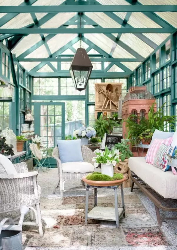a she shed in vintage style of a former glasshouse, with white wicker furniture, tables, a sofa, potted plants and some vintage furniture