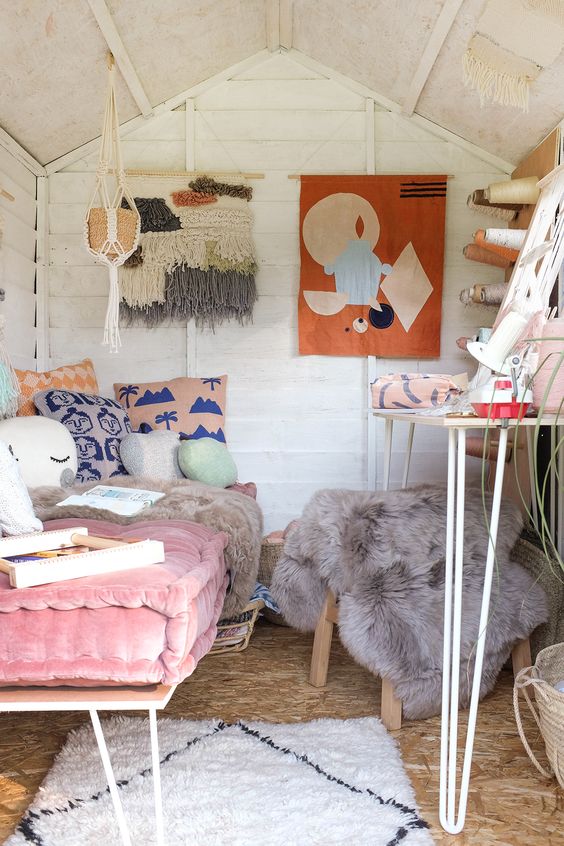 a small and lovely boho she shed with a crafting space with a desk, a pink daybed with lots of pillows and boho art