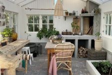a small and lovely she shed with a hearth, a wooden table for potting, a wooden table and rattan chairs, potted plants