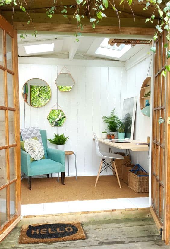 a small and lovely she shed with white planked walls, a floating desk, a white chair, a mint one, potted greenery and mirrors