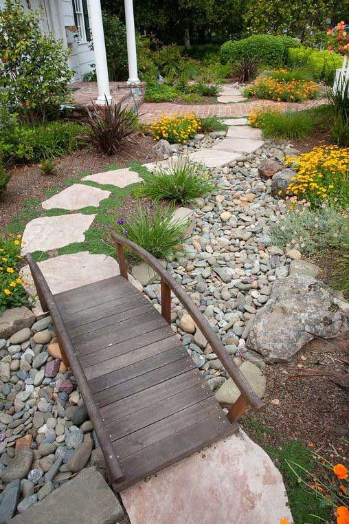 a small water wise garden with pebbles and rocks, bright blooms and greenery plus a stained bridge over the rocks for an eye catchy touch