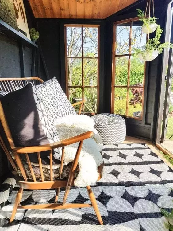 a small yet lovely modern she shed with a wooden daybed with pillows, potted greenery, a pouf and a printed rug