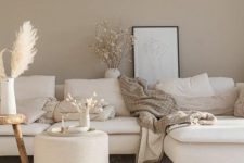 a soothing greige living room with a creamy sectional and pillows, a creamy pouf and a wooden side table plus some decor