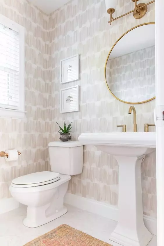 a stylish neutral powder room with printed wallpaper, a round mirror in a gilded frame, a free-standing sink and some plants
