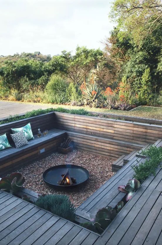 a sunken conversation pit with gravel on the ground, a fire pit, built-in wooden benches around and some growing greenery