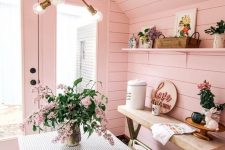 a super cute she shed with pink planked walls and a ceiling, a bench, an open shelf, an upholstered able and chairs and some blooms