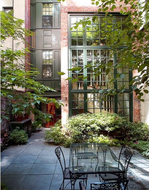 a townhouse courtyard done with stone tiles, metal chairs and a glass table and some greenery and trees around
