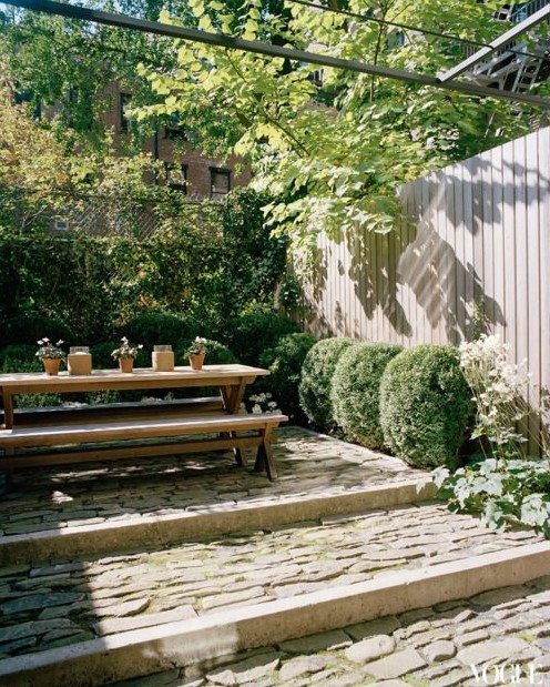 a townhouse garden with stone steps, greenery and boxwood growing and a simple wooden dining set with benches