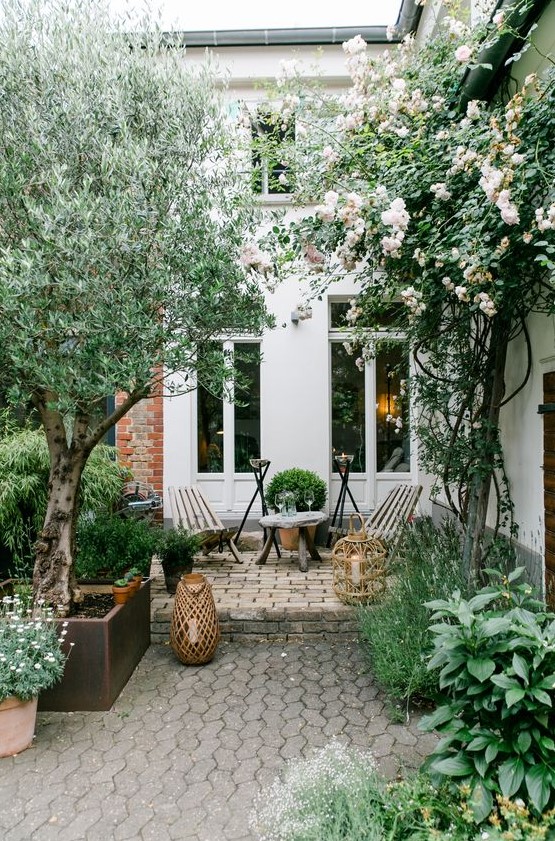 a townhouse garden with stone tiles and bricks, trees, shrubs and greenery, simple wooden furniture and candle lanterns