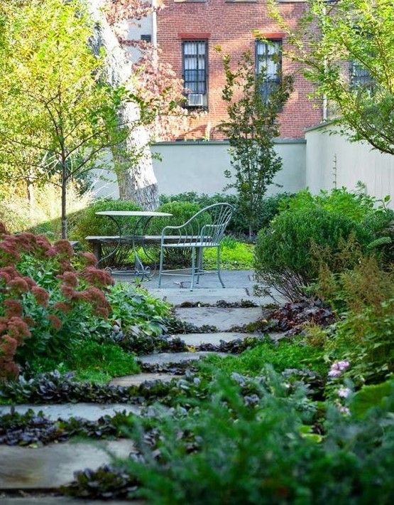 a townhouse garden with trees and lush greenery growing even in between the stone steps and metal furniture
