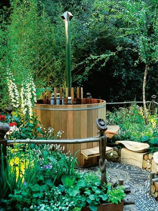 a whimsical hot tub space in the garden with lots of greenery and blooms, with a mosaic walkway and a wooden bathtub with a step