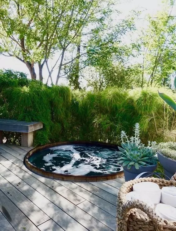 a wooden deck with a built in hot tub, surrounded with a greenery wall, with a bench, potted plants and baskets with towels