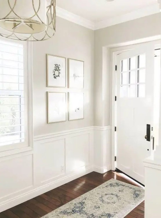 an airy and light-filled greige entryway with white wainscoting, a chandelier, a mini gallery wall and a white door