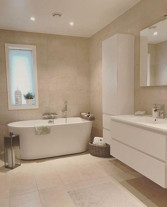 an eclectic greige bathroom clad with large scale tiles, a white storage unit and a vanity, a white tub and a large lantern