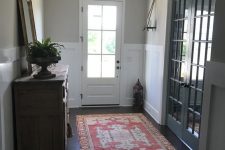 an eclectic greige entryway with white paneling, a stained dresser, a bold red rug, a mirror in a chic frame and greenery
