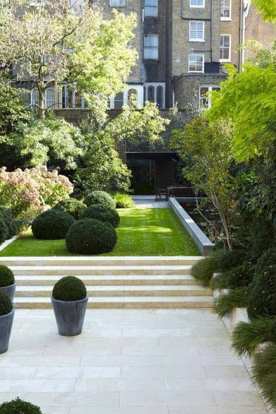an elegant minimalist townhouse garden with stone tiles and steps, a lawn, boxwood, trees and shrubs plus a flower bed