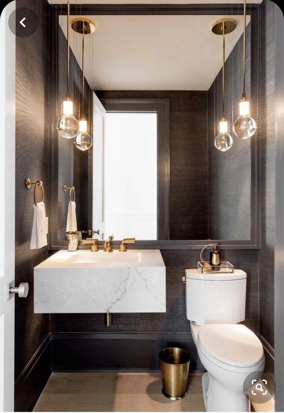 an elegant modern powder room with black walls, a white stone slab sink and a white toilet, pendant bulbs and a large mirror