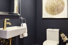 an elegant modern powder room with navy walls and a black hex tile floor, a wall-mounted sink and a toilet, gold touches for more chic