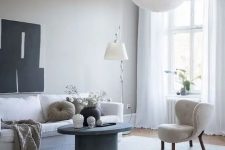 an ethereal greige living room with a white sofa and a cool wingback chair, a grey concrete round table, a floor and a pendant lamp