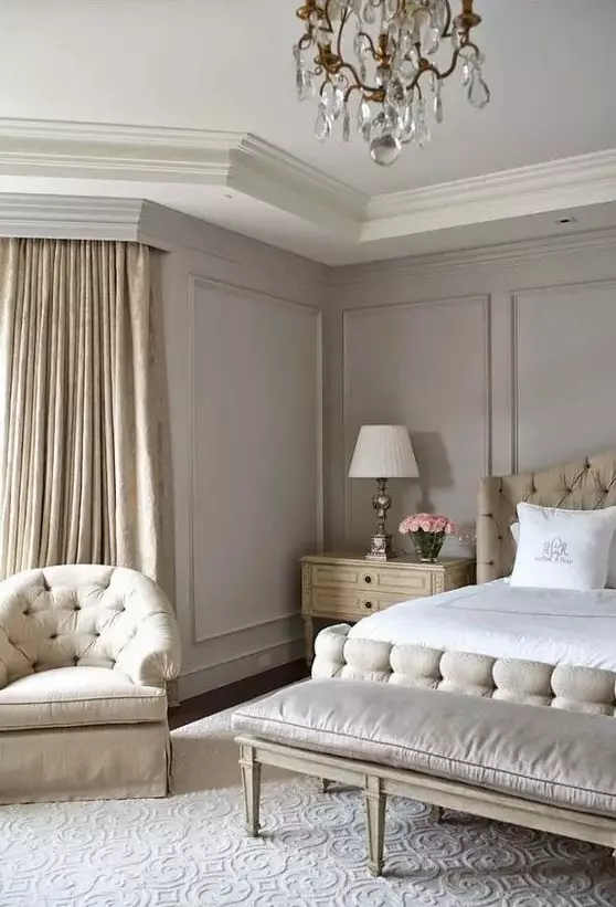 an exquisite greige bedroom with neutral upholstered furniture, a bench, neutral bedding, a crystal chandelier and tan printed curtains