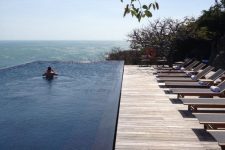 an infinity pool with a fantastic sea view, a simple wooden deck and a whole row of minimalist loungers and trees around