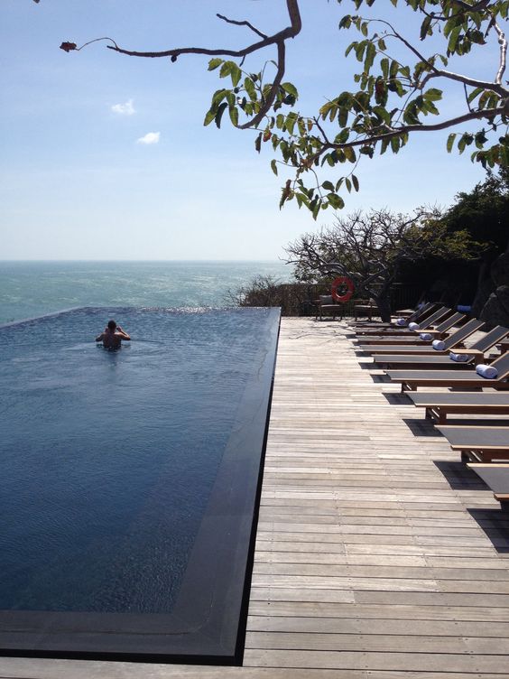 an infinity pool with a fantastic sea view, a simple wooden deck and a whole row of minimalist loungers and trees around