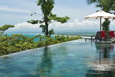 an infinity pool with greenery around and a sea view, with some outdoor furniture is a cool space to spend a hot day