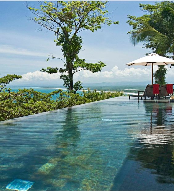 an infinity pool with greenery around and a sea view, with some outdoor furniture is a cool space to spend a hot day