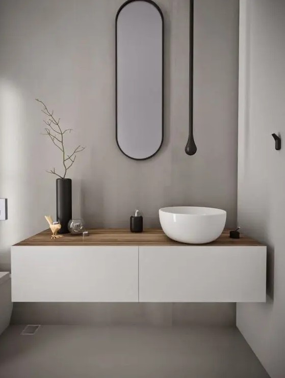 an ultra-minimalist powder room with grey walls, a floating white vanity with a bowl sink, a long and narrow oval mirror, a black light and a toilet