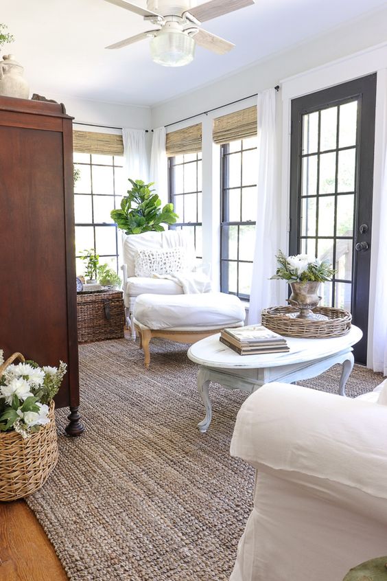 a French farmhouse sunroom with a jute rug, a stained wardrobe, white chairs and a coffee table, potted blooms and plants