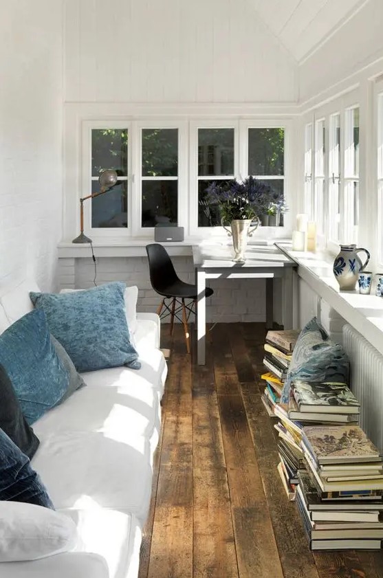 a Nordic sunroom and a home office in one with a windowsill desk a white sofa with blue pillows lots of books and some blooms