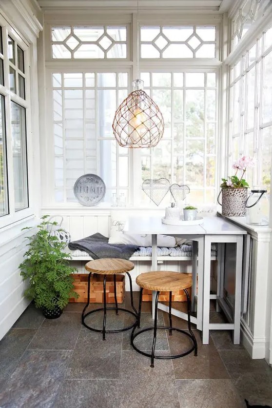a Nordic sunroom in white, with grey tiles on the floor, a built-in bench, a white table, wooden stools and a woven pendant lamp
