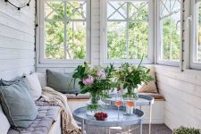 a Scandinavian sunroom with a series of windows, a built-in bench, pastel upholstery and pillows, round coffee tables and potted blooms