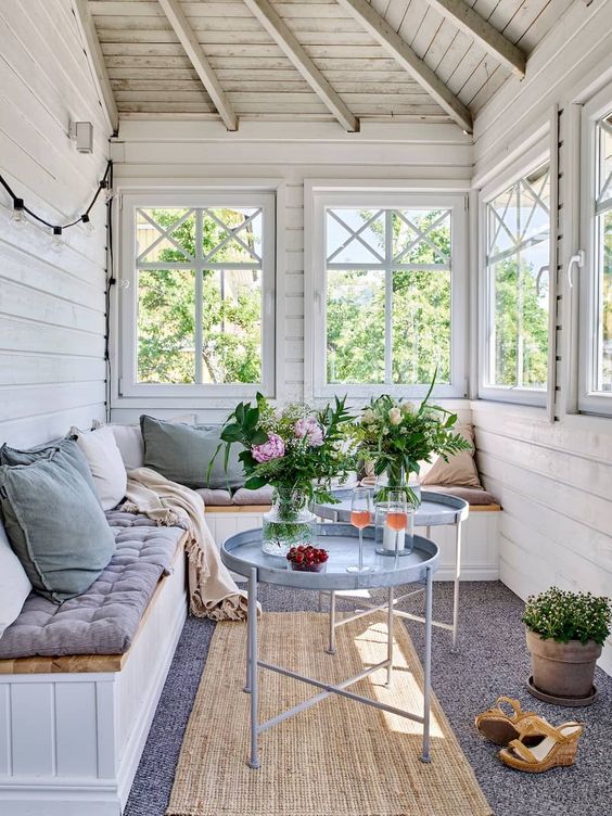 a Scandinavian sunroom with a series of windows, a built-in bench, pastel upholstery and pillows, round coffee tables and potted blooms