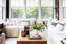 a beautiful Scandinavian sunroom with a white built-in bench with neutral upholstery and printed pillows, a low coffee table, a potted plant and a rattan chair