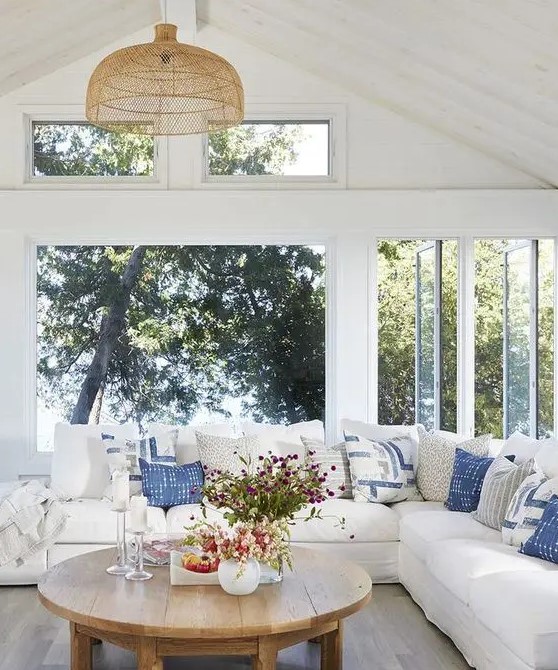 a beautiful modern farmhouse coastal sunroom with a large sectional, blue pillows, a wooden table and woven chandelier