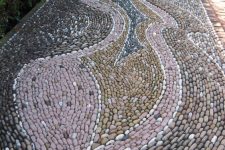 a beautiful mosaic pebble garden path with pink, mustard and grey patterns and greenery along the path is amazing