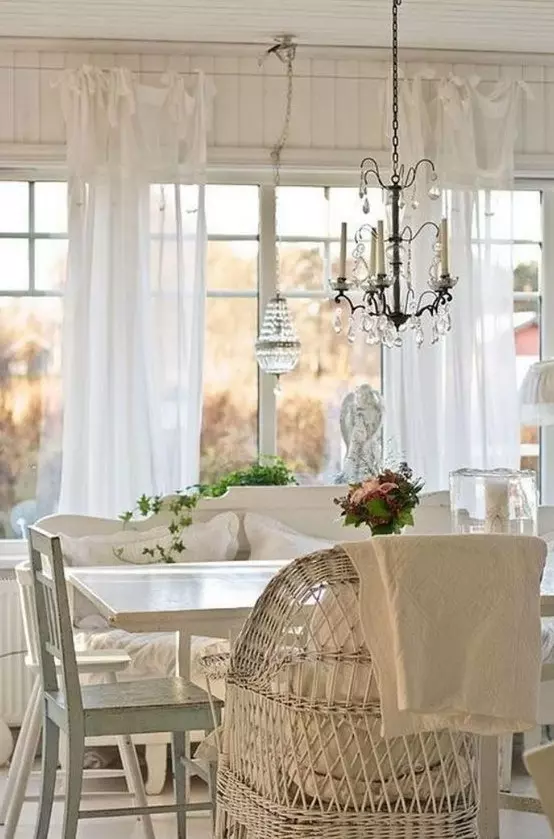 a beautiful vintage cottage sunroom in white, with wooden and wicker furniture, a crystal chandelier and greenery