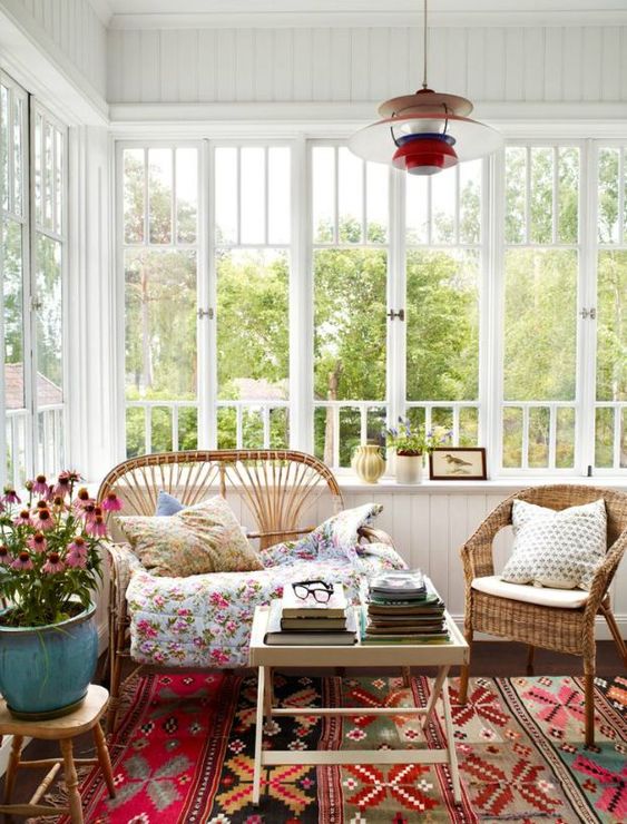 a boho sunroom with a touch of vintage, with rattan and wicker furniture, a bold printed rug and blankets, potted blooms and a colorful lamp