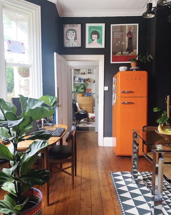 a bold and catchy space with navy walls, an orange fridge, a stained desk and a black chair, a bold mini gallery wall with catchy artwork