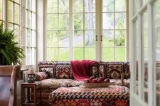 a bold boho sunroom with a red printed boho sofa, a matching ottoman, a red printed rug, rich-stained furniture and potted greenery