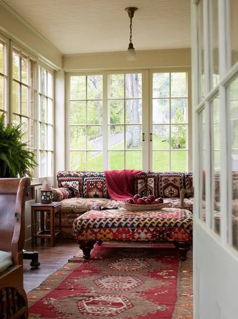 a bold boho sunroom with a red printed boho sofa, a matching ottoman, a red printed rug, rich stained furniture and potted greenery