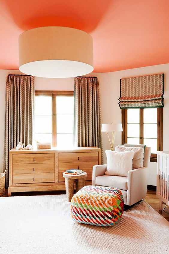 a bold nursery with creamy walls and a coral ceiling, a stained dresser, a white chair, a colorful pouf and curtains
