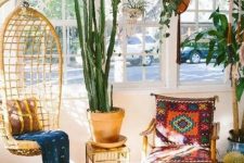a bright boho sunroom space with a wicker pendant chair, a colorful chair, layered rugs, potted plants and a view