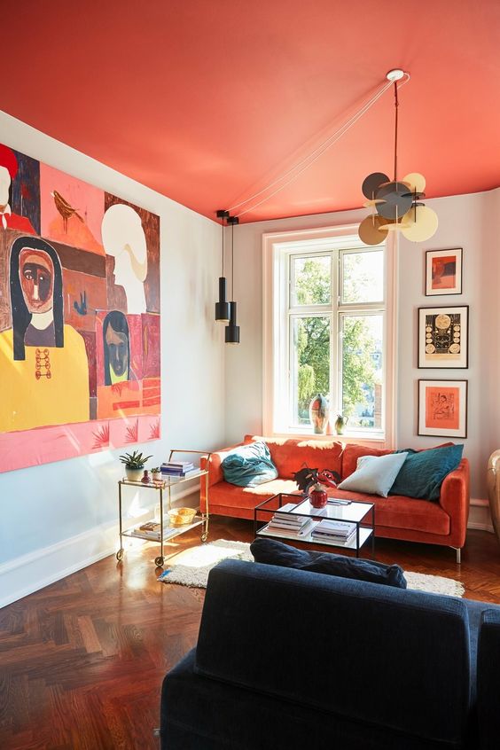 a bright living room with a hot red ceiling, an orange sofa, a navy chair, a bold artwork and pendant lamps plus elegant coffee tables