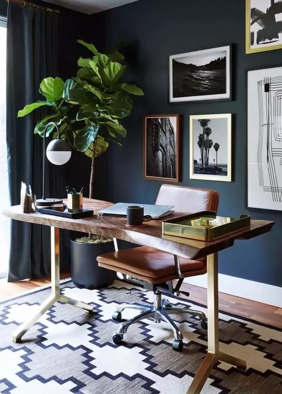 a chic mid-century modern home office with navy walls and curtains, a living edge desk, a leather chair, a catchy gallery wall and a potted tree