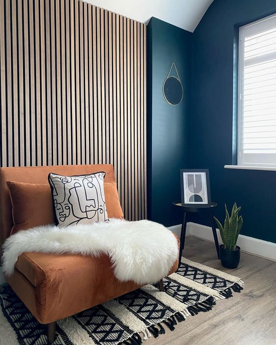 a chic mid-century modern nook with a wood slat accent wall, a rust-colored chair with pillows, a printed rug, a side table and a potted plant
