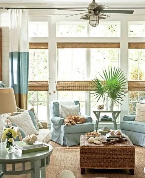 a coastal sunroom with neutral and light blue furniture, color block curtains, woven shades and a wicker table plus greenery