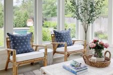 a coastal sunroom with rattan chairs and blue pillows, a white wicker table and printed textiles, a wicker lamp looks chic