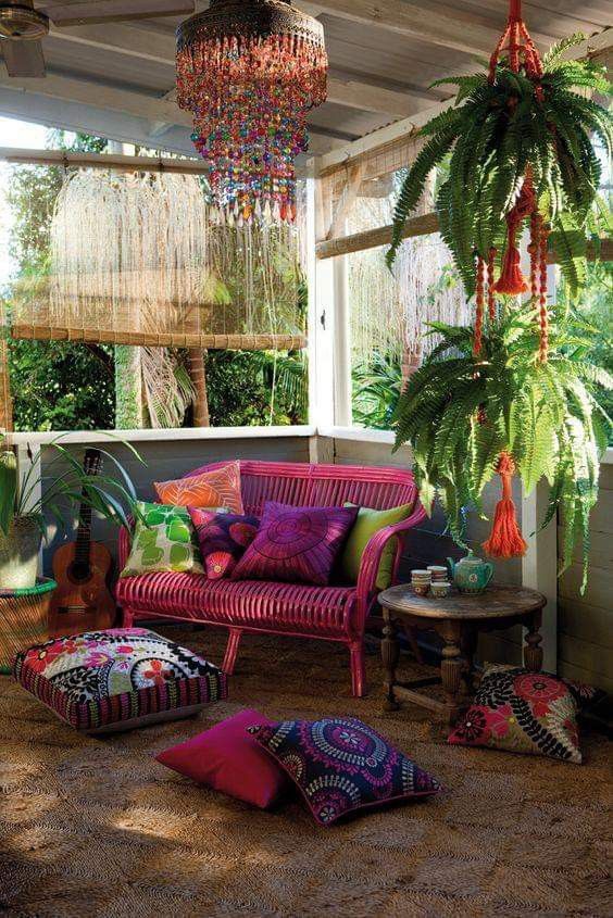 a colorful boho sunroom with a hot pink loveseat, bold pillows on the floor, potted plants, a colorful chandelier and a side table